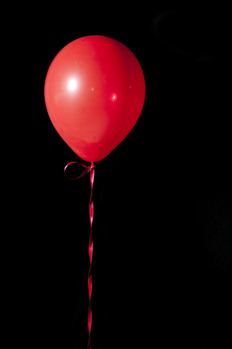 a single red balloon tied with ribbon floating in the air against a black backdrop