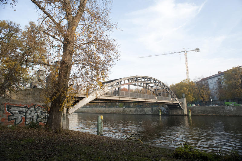 Arched bridge over the river Spree in Berlin with a tree on the bank in the foreground and an industrial crane visible in the distance on the horizon