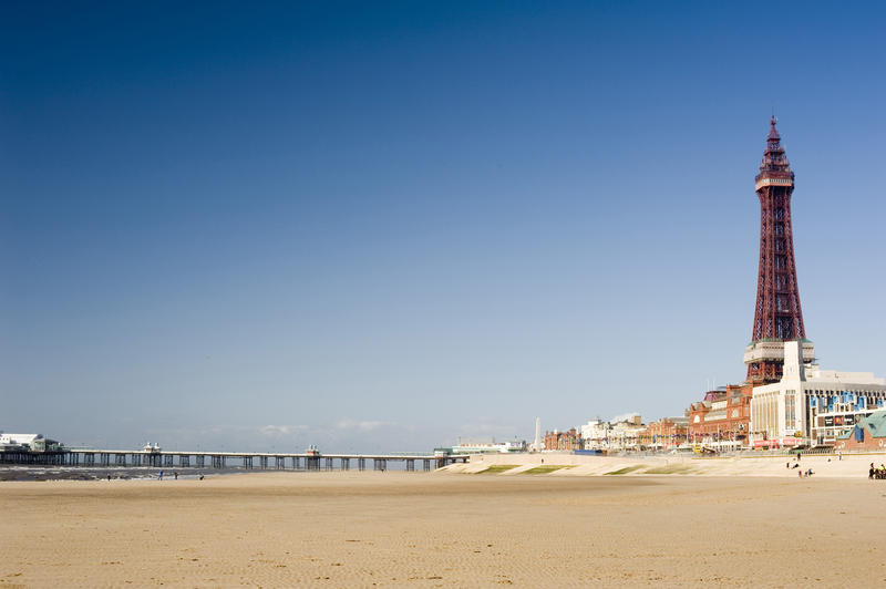 Open expanse of sand at the Blackpool beach with the Blackpool Tower and waterfront on the right and central pier in the distance