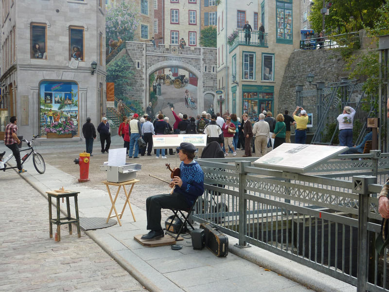 Busker in Quebec City sitting on the sidewalk watching a crowd of people admiring a trompe d'oeil painting on a building facade