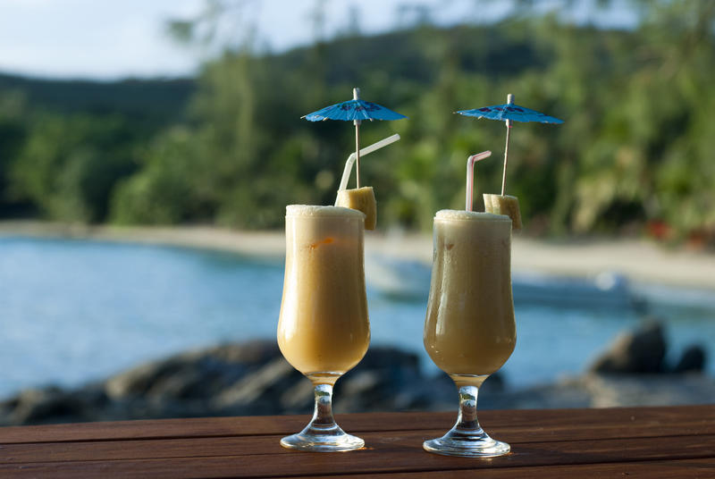 Two celebratory refreshing tropical cocktails served with small cocktail umbrellas on a tabletop overlooking an idyllic bay and beach on summer vacation