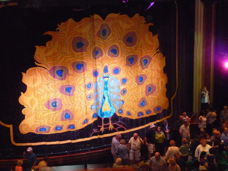 People gathering for a performance on a cruise ship in front of the stage in the theatre which is enclosed by a decorative curtain with a peacock