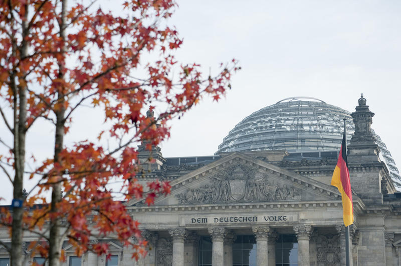 View past colourful red autumn leaves on a tree to the Reichstag building , Berlin showing the dedication to the people on the architrave and the glass dome