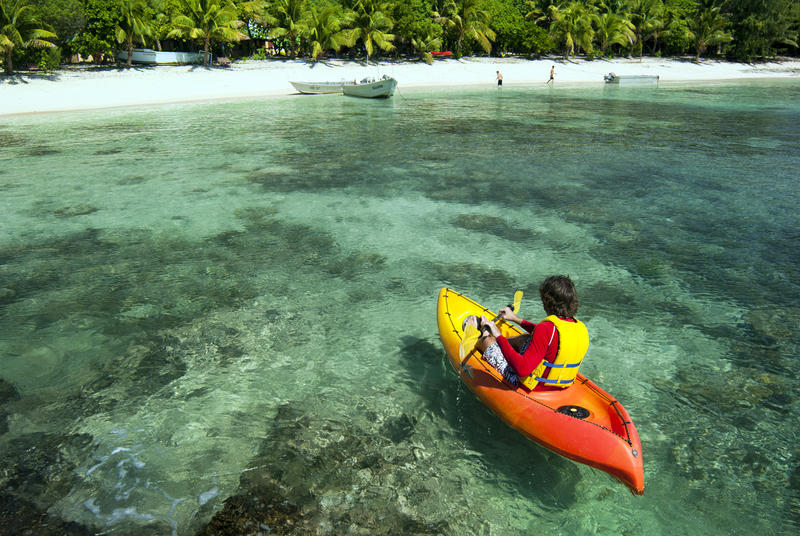 Kayaker paddling towards a sandy beach over crystal clear tropical water with shallow ocean rocks visible below