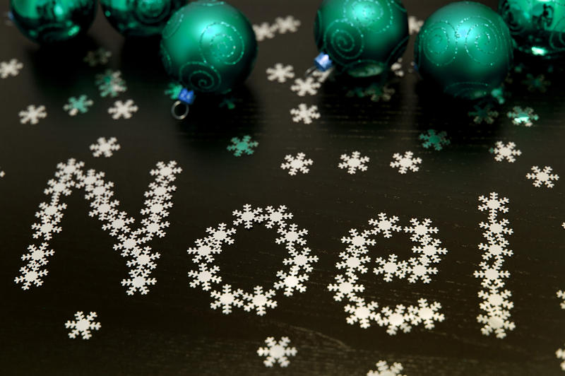 Noel snowflake greeting with tiny snowflake decorations forming the word Noel on a black background amidst scattered green baubles