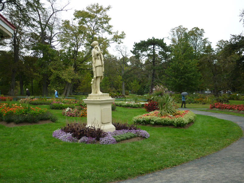 Statue in a park with pretty colourful flowerbeds, green lawns and walkways with a person taking an afternoon stroll in the distance