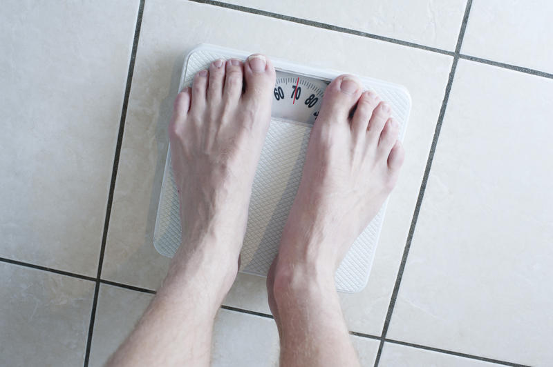 Overhead view of a barefoot man weighing himself on a bathroom scale to monitor his weight gain or loss