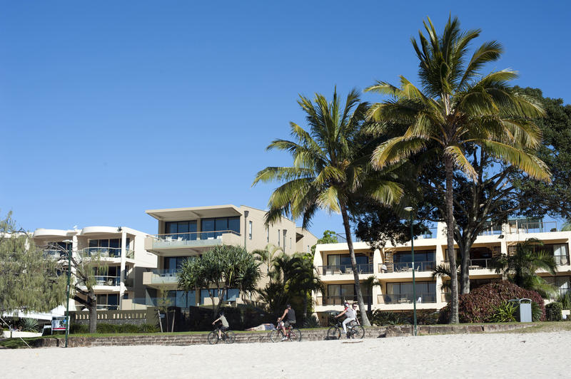 Modern tropical beachfront hotel with people riding bicycles in the foreground and palm trees against a sunny blue summer sky