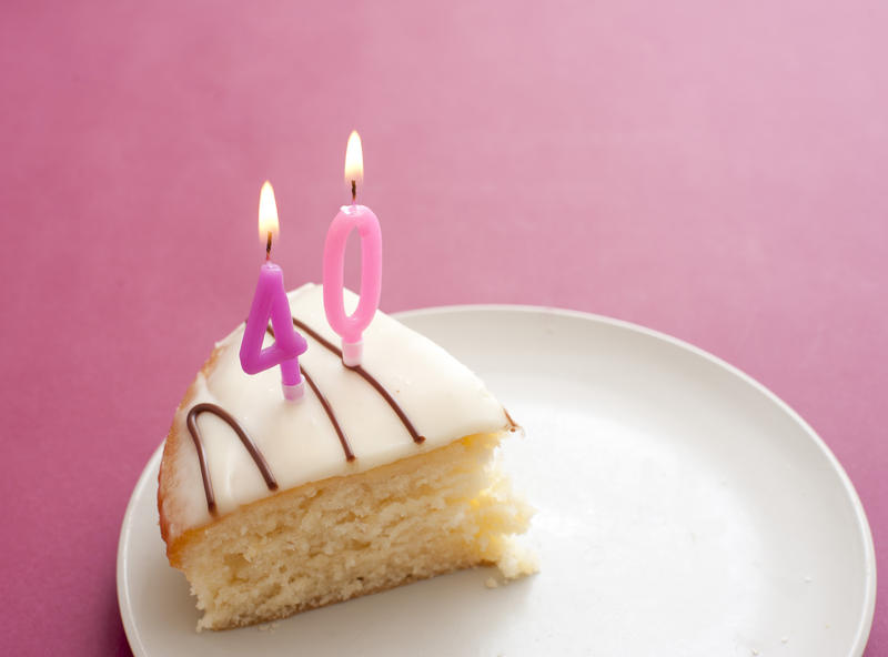 40th Birthday Cake with two pink number candles burning on a slice of iced cake served on a plate over a matching pink background celebrating the birthday of a lady or woman