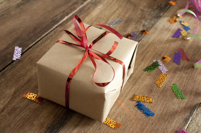 Brown Birthday Gift Box with Red Ribbon Placed on a Wooden Table with Confetti.