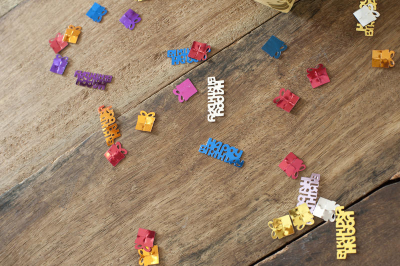 Colorful Birthday sprinkles or confetti with Happy Birthday text or multicolored gifts shapes tied with ribbon scattered randomly on a wooden table with copyspace for your greeting