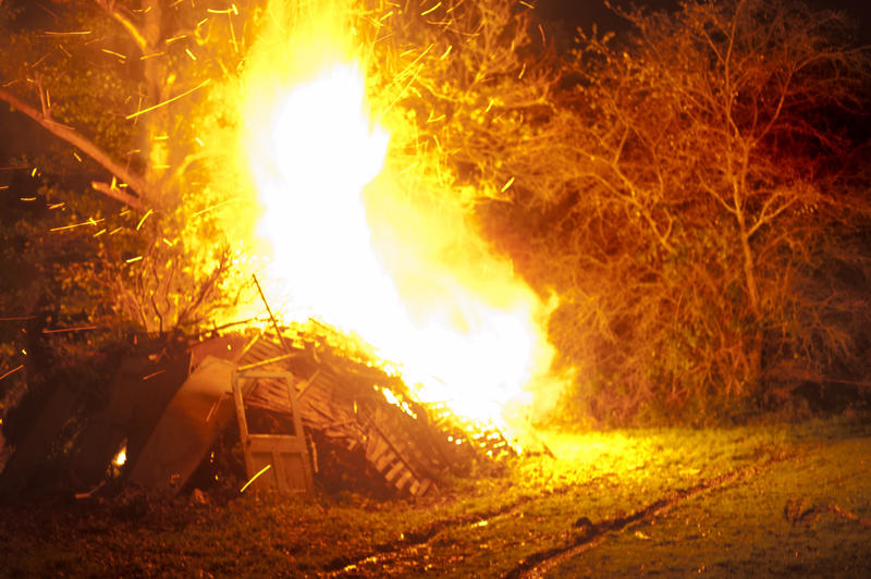 Roaring bonfire with bright orange flames shooting into the air in a rural garden or field lighting up the trees in the darkness