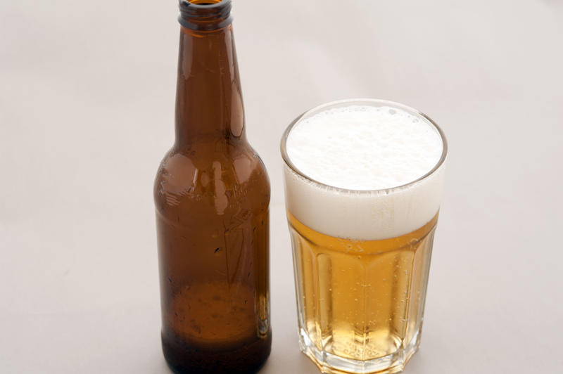 Bottled beer in a tall glass with a frothy head with the almost empty unlabeled brown bottle standing alongside, over grey