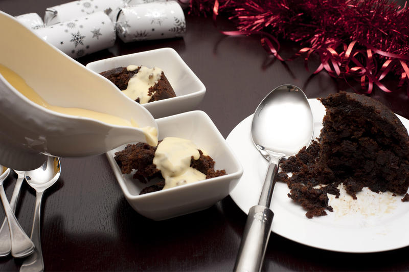 Pouring brandy custard onto servings of delicious fruity Christmas pudding on a festive table with ornaments and Xmas crackers