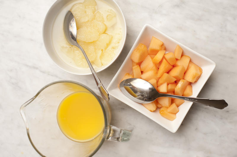 Variety of breakfast fruits with orange juice in a jug, and fresh diced melon and pears in individual dishes, overhead view
