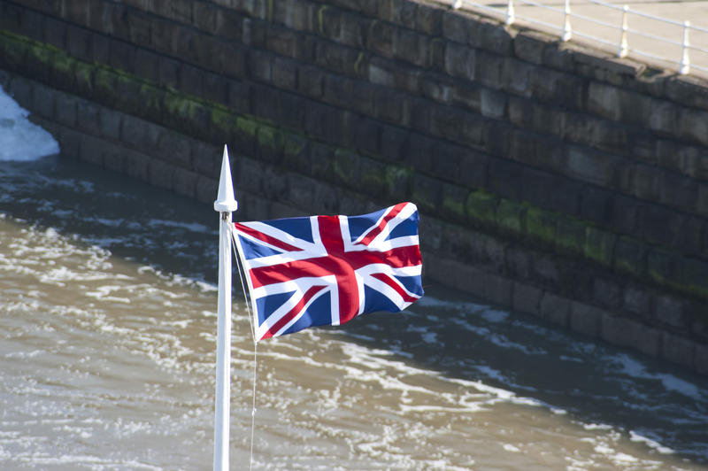 British national flag, the Union Jack, flying from a flagpole in front of a coastal pier with foamy sea water