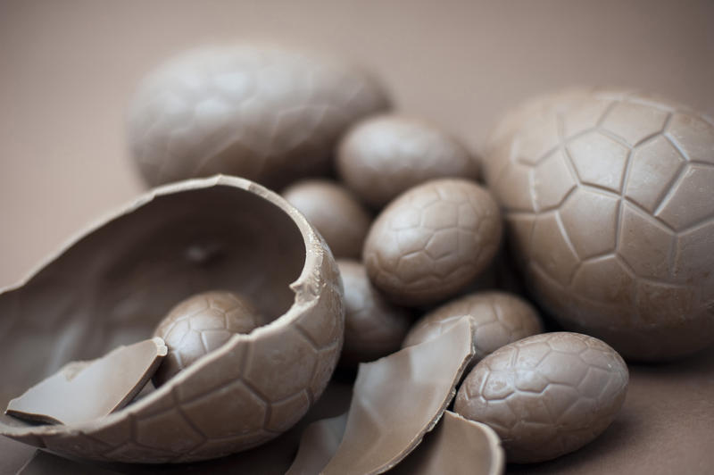 Brown background with a broken chocolate Easter egg in the foreground and an assortment of whole eggs behind with shallow dof