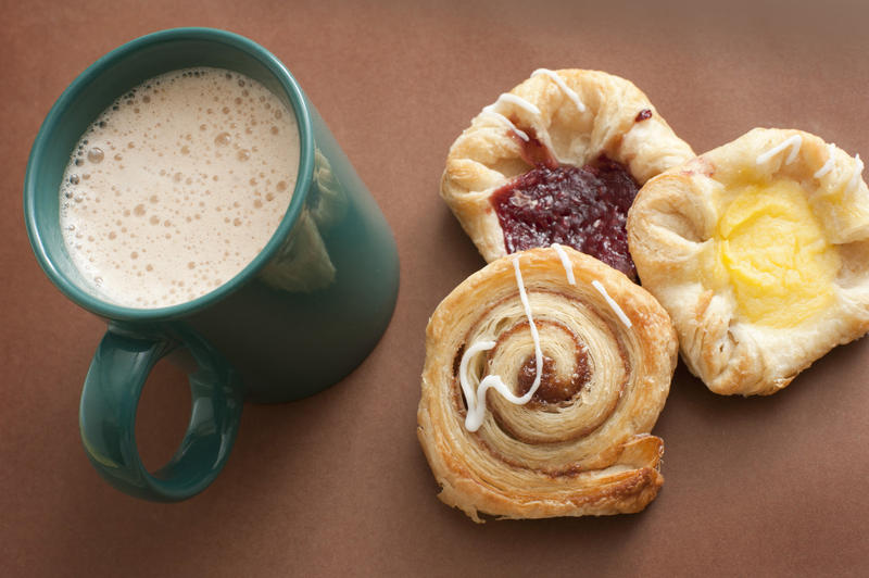 Cup of freshly brewed frothy cappuccino with assorted pastries including a cinnamon Danish, custard tart and jam pastry, view from above