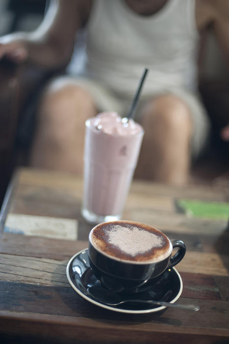 Cup of cappuccino and a milkshake on a table in a restaurant or cafeteria with a persons legs visible behind