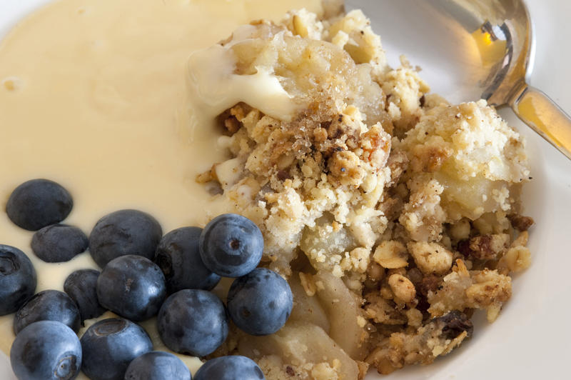 Close up view of a bowl of delicious apple crumble topped with spicy raisins in sugar and served with whole fresh blackcurrants and custard