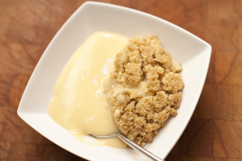 Tasty apple crumble dessert served with custard in a modern square pudding bowl on a wooden table