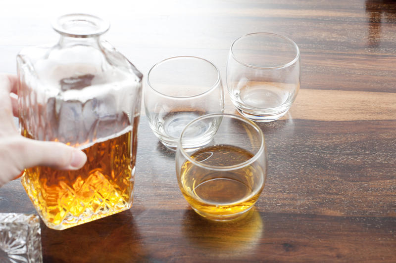 Man serving whiskey or brandy from a square glass decanter into circular glasses on a wooden bar counter, close up of his hand