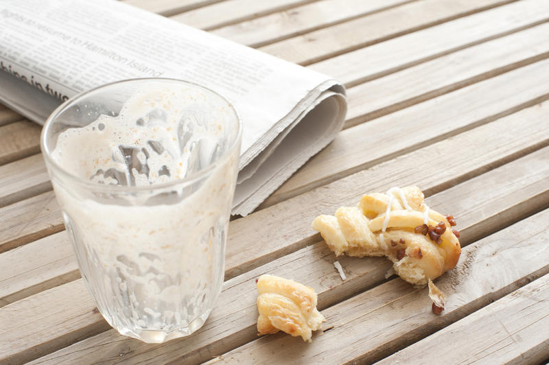Close Up of Empty Coffee Glass and Half Eaten Pastry on Wooden Table Next to Rolled Newspaper - Remnants of Breakfast or Coffee Break on Wood Patio Table