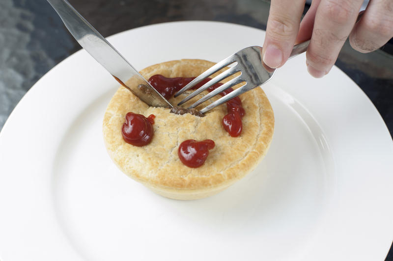 Man eating a freshly baked golden meat pie for lunch with a smiling gravy face on the pastry crust, high angle view as he cuts into it with a knife on a white plate