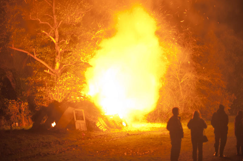 People standing watching a blazing bonfire as fiery orange flames leap into the air lighting up the trees behind on Guy fawkes or Bonfire Night