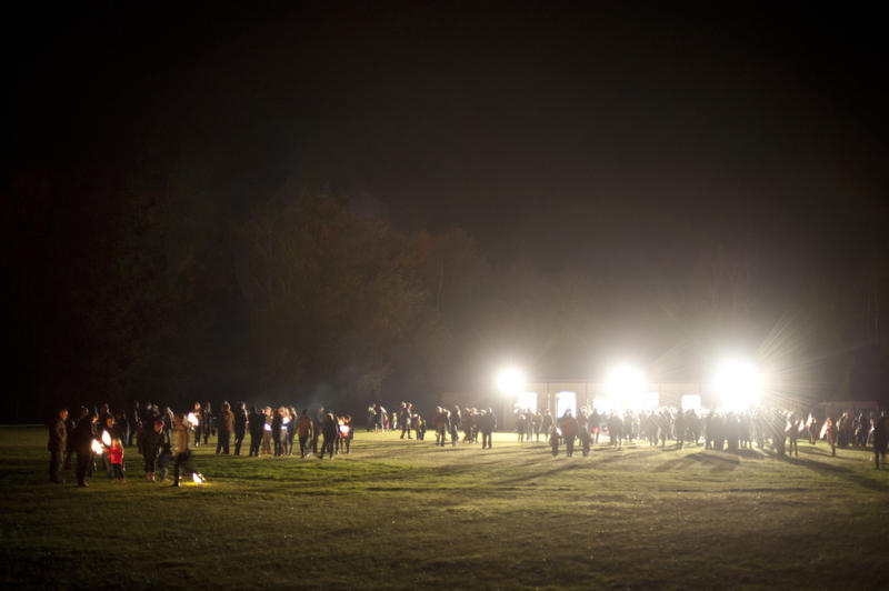 Three bright floodlights lighting a festival with crowds of people gathered on a field to celebrate Bonfire Night on the 5th November