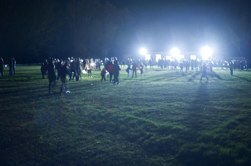 Floodlit night event with crowds of people walking across an open field illuminated by a row of powerful floodlights facing towards the camera in the darkness