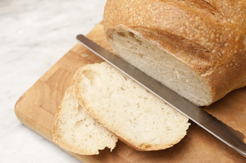 Slicing a loaf of white bread with two slices and a knife with a round loaf on a wooden bread board over white