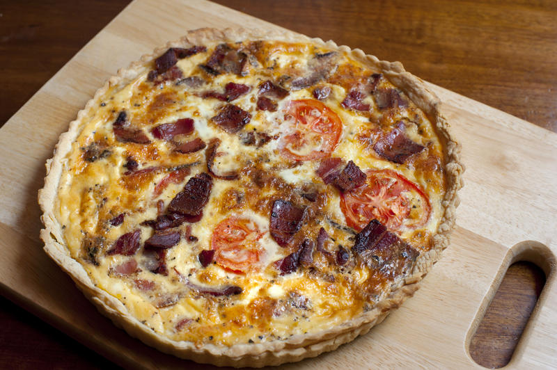 Homebaked savoury egg quiche seaoned with bacon, cheese and tomato in a crisp pie crust standing on a wooden chopping board