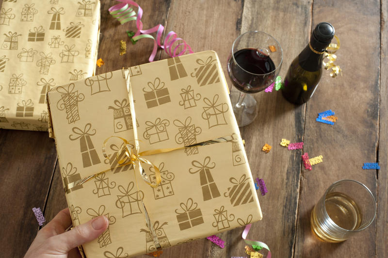 High Angle View of a Hand Holding a Yellow Gift Box on Top of a Wooden Table with Wine, Streamers and Confetti.