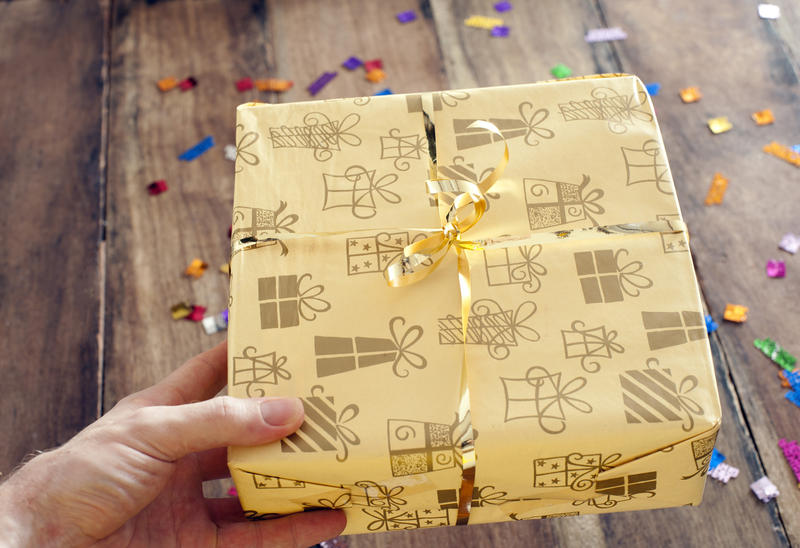 Man giving a decorative birthday gift in pretty patterned gift wrap holding out his hand above a wooden table with colorful party sprinkles