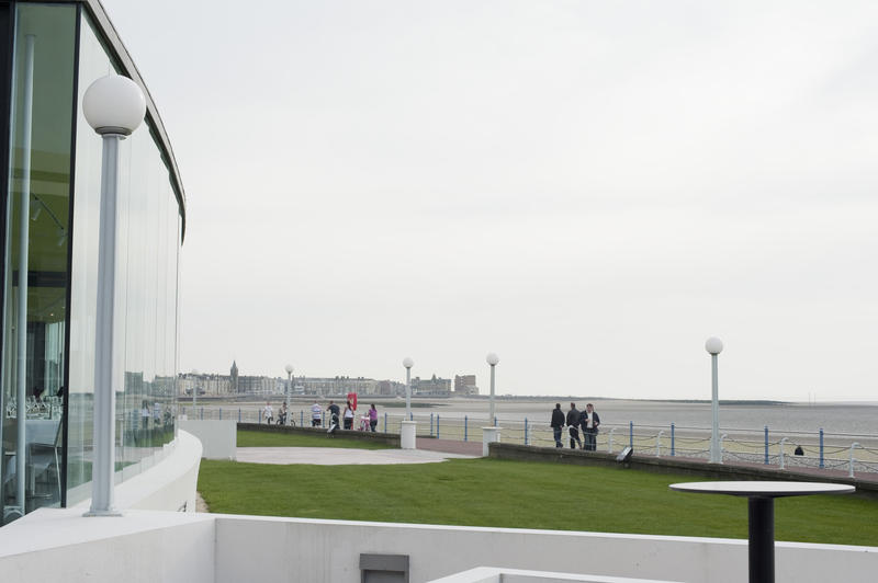 Morecambe promenade and the front facade of the iconic Art Deco Midland Hotel building with people walking along the seafront