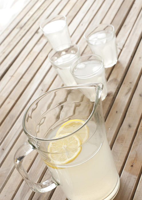 Fresh homemade tangy lemonade with slices of lemon in a glass jug and tumblers on a wooden picnic table in summer