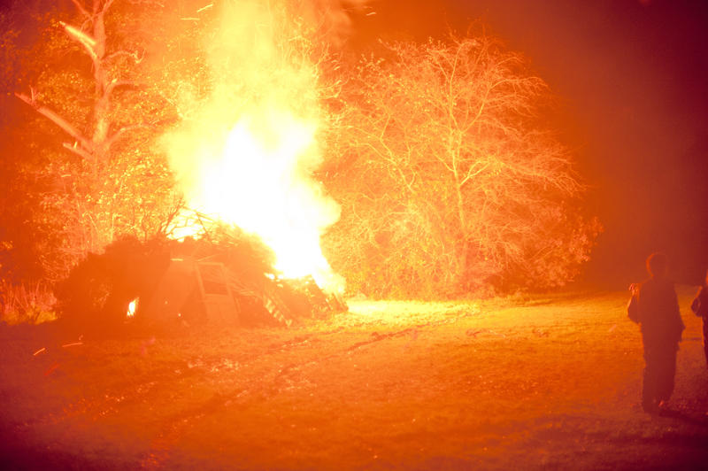 Blazing hot bonfire with fiery orange flames shooting into the air illuminating the surrounding darkness and trees on the field with a few people watching from the right side as it burns
