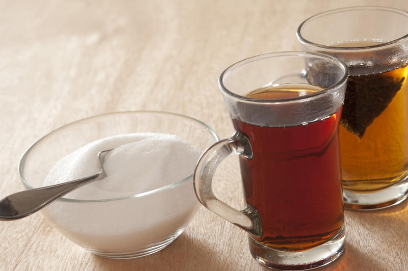 Two glass mugs of hot sweet black tea, one with a teabag still steeping, alongside a bowl of white sugar