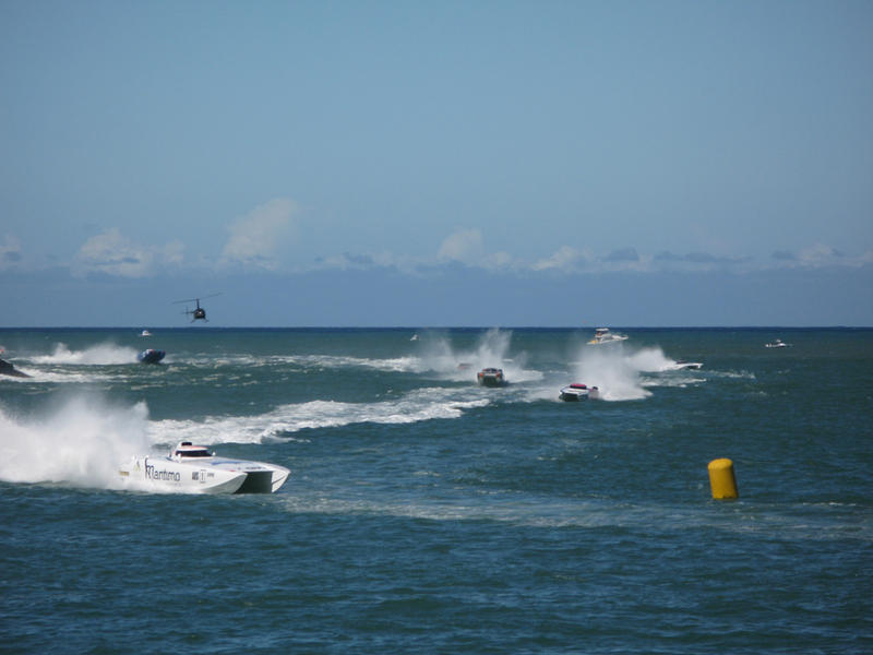 Jet boats race with speeding boats rounding a marker buoy offshore in the ocean throwing up spray in their wakes with a helicopter hovering above