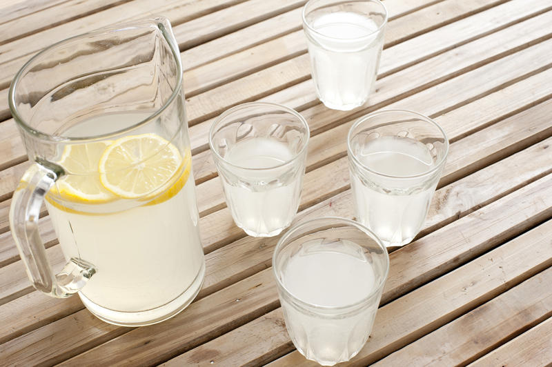 Delicious refreshing homemade lemonade with slices of fresh lemon served in a glass jug and tumblers on a wooden slatted picnic table in summer