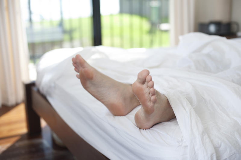 Man having a weekend lie-in in his bed with a view of his feet sticking out of the end of the white linen bedclothes and a window in the background