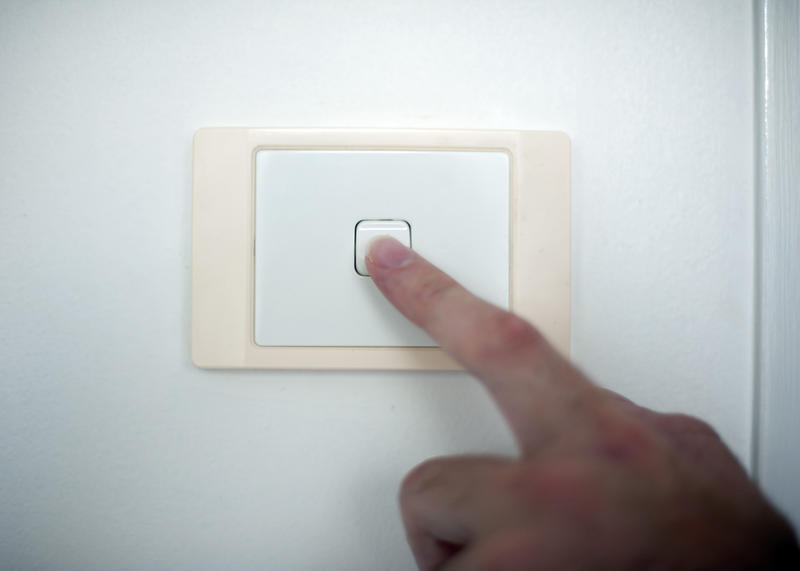 Finger switching on or off a white plastic wall-mounted light switch in a close up conceptual view of power, energy and electricity consumption and supply