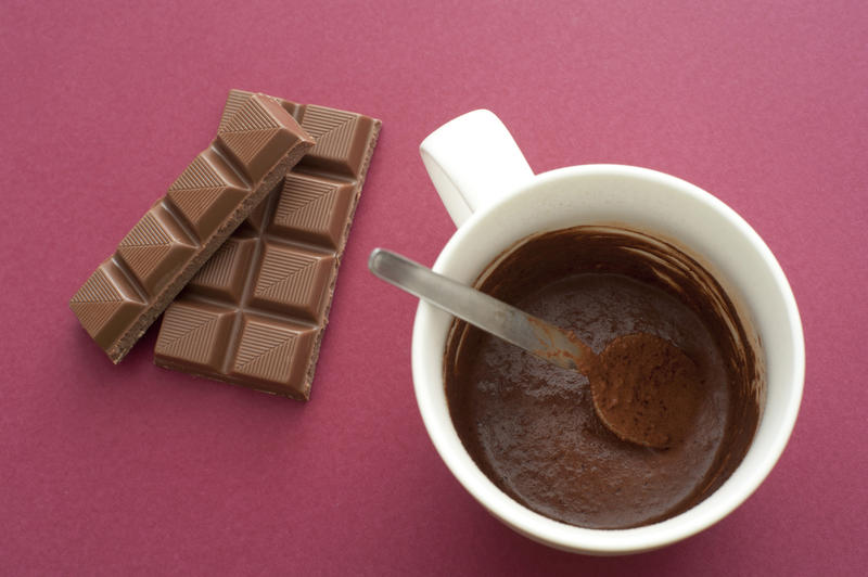 High Angle View of Milk Chocolate Candy Bar Squares Next to Mug of Melted Cocoa with Spoon on Dark Pink Background with Copy Space