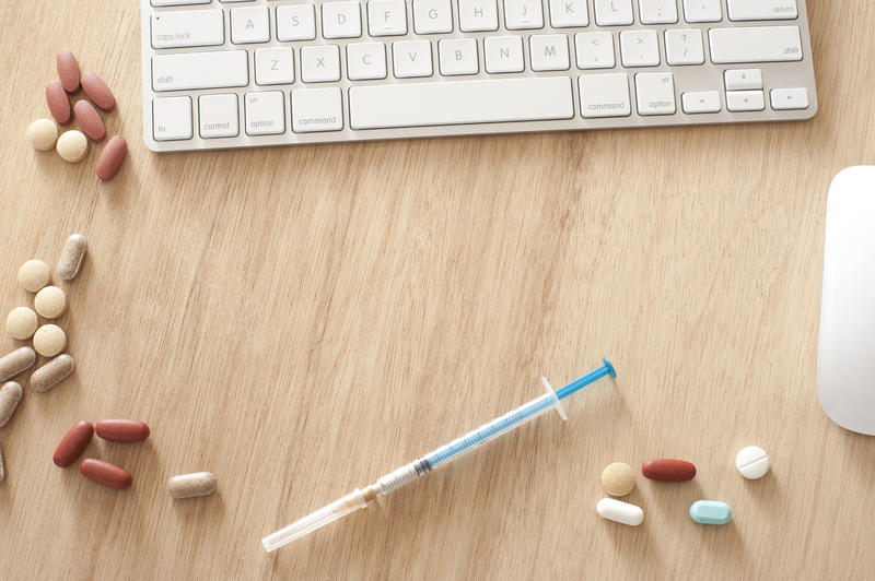 Online medical concept with a computer keyboard, mouse, assorted pills and tablets and a syringe and needle on a wooden table with central copyspace