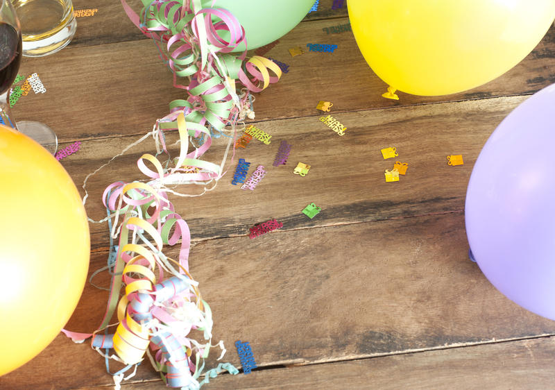 Festive party backdrop with an overhead view of coiled streamers and confetti on a wooden table surrounded by a frame of colorful balloons, with copyspace