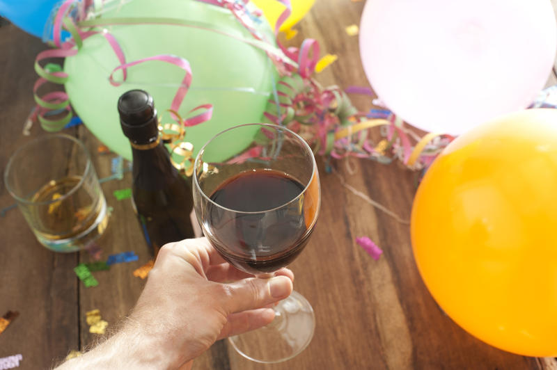 Man drinking red wine at a party holding his glass above a glass of spirits and bottle of champagne on background of colorful party balloons and streamers in a celebration concept