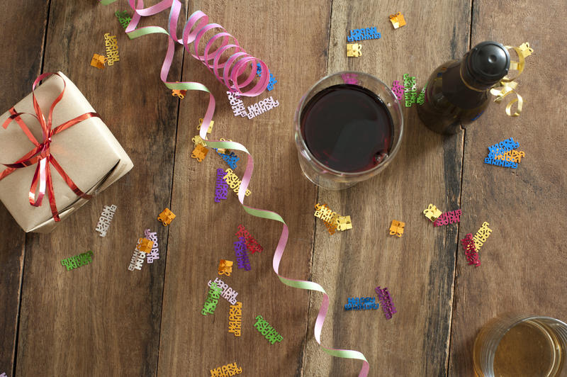 Overhead view of a gift-wrapped party gift and drinks with a bottle of champagne and glass of red wine with scattered colorful streamers and confetti