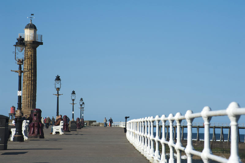 View along the West Pier at Whitby showing people walking on the promenade with the navigation lighthouse at the end