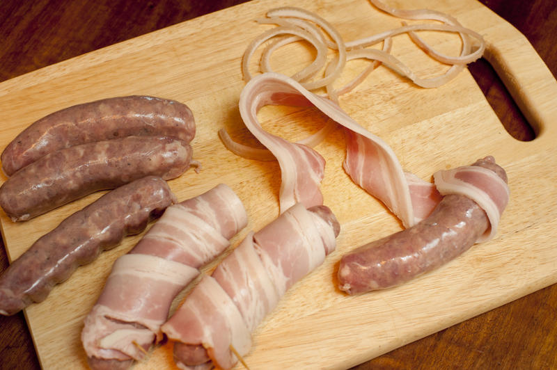 Preparing Pigs In Blankets or sausages wrapped in rashers of fresh smoked bacon with the ingredients laid out on a wooden chopping board